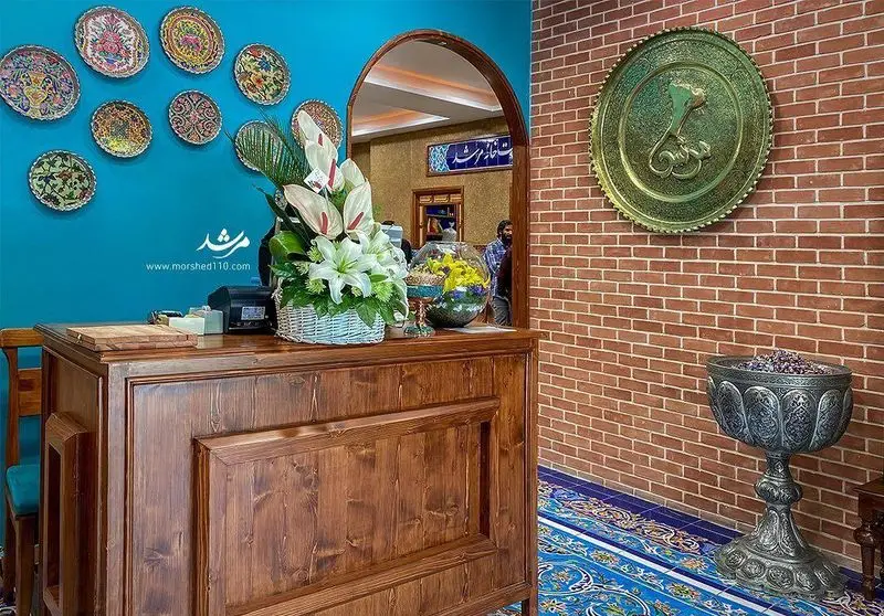 فضای داخلی رستوران سنتی مرشد باهنر، شامل میز پذیرش چوبی، دیوار آبی با بشقاب‌های دکوراتیو و دیوار آجری با آینه قوسی و ظروف قدیمی.