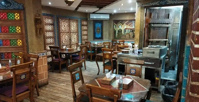 فضای داخلی دلنشین و دکوراسیون سنتی رستوران مرشد باهنر، با میز و صندلی‌های چوبی، پنجره‌های طرح‌دار و نقاشی‌های دیواری اصیل ایرانی.