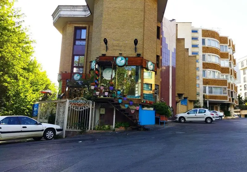نمایی بیرونی از رستوران مرشد باهنر با نمای آجری، گلدان‌های رنگی، درختان سبز و خودروهای پارک شده در خیابان، تهران.