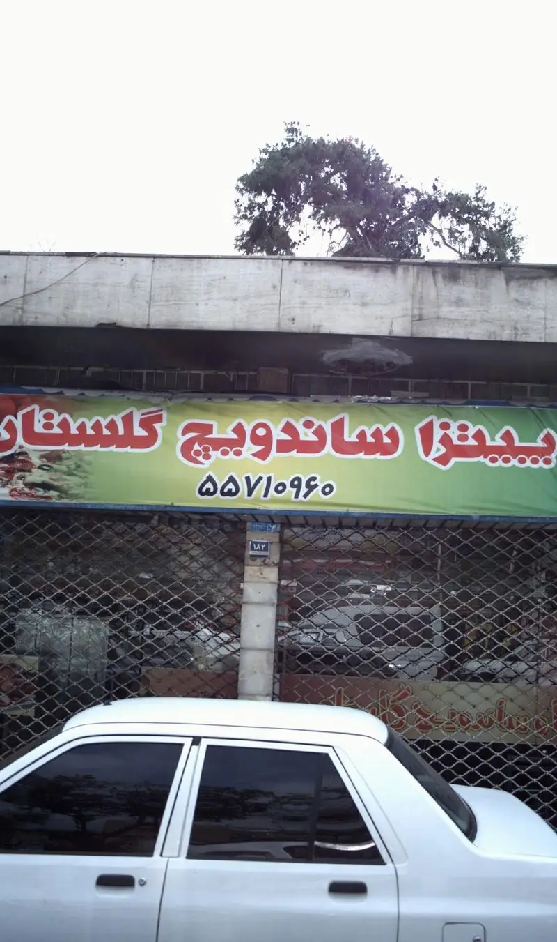 تصویر تابلوی سبز رنگ پیتزا ساندویچ گلستان با فونت قرمز و شماره تماس ۵۵۷۱۰۹۶۰. نمای بیرونی مغازه بسته با کرکره فلزی.