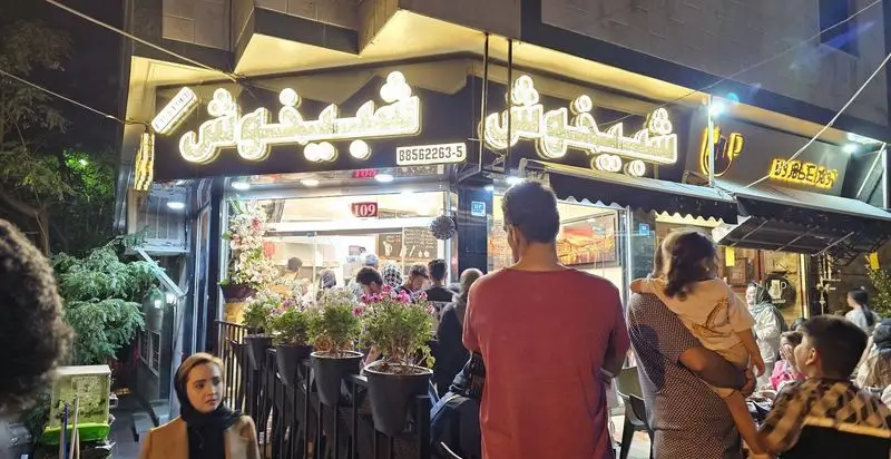 تصویری از نمای شبانه و پر جنب و جوش فست فود شبیخوش، با تابلوهای نئون روشن و جمعیتی از مشتریان در حال انتظار.