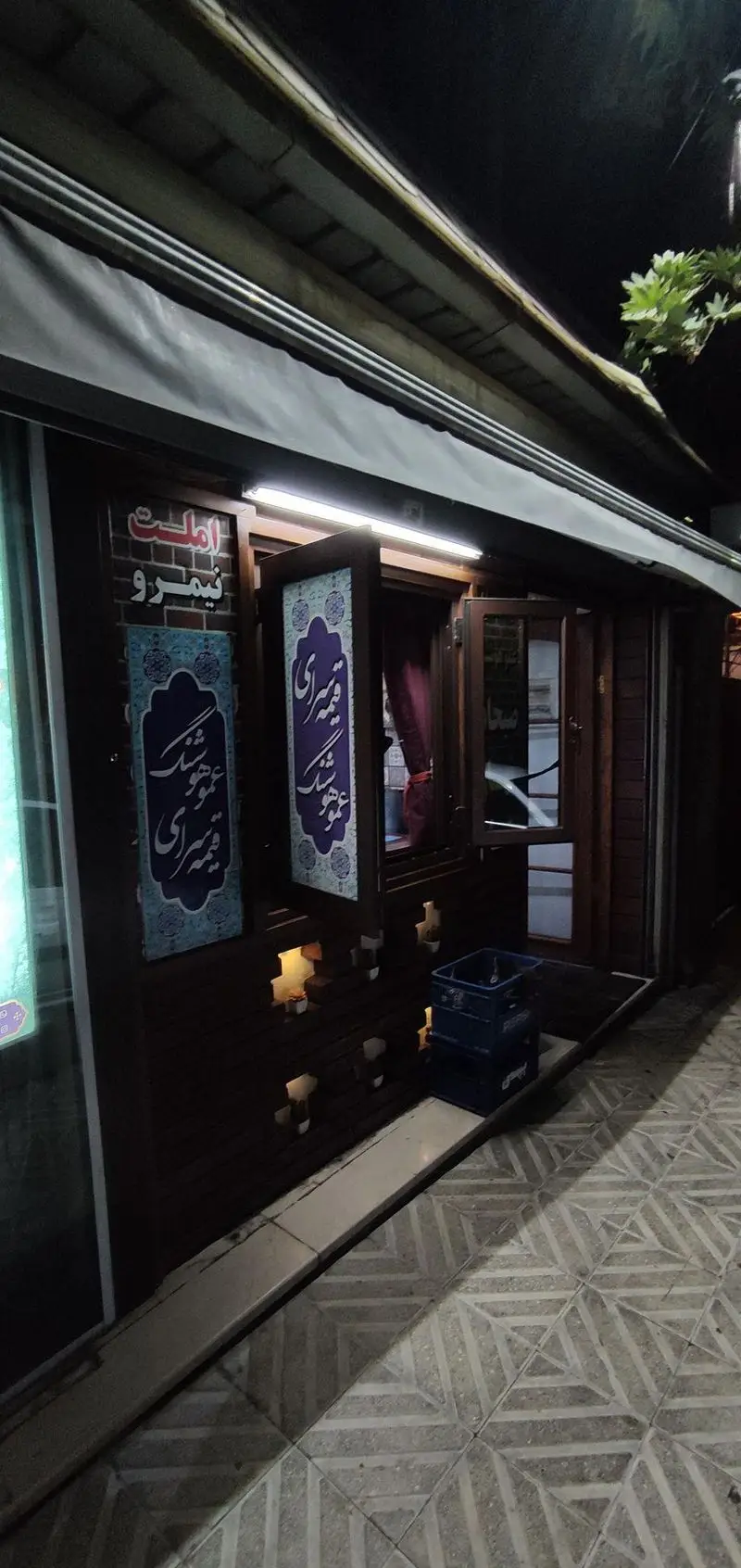 نمای بیرونی قیمه سرای عمو هوشنگ در شب با سردر چوبی، سایبان، دو بنر کالیگرافی فارسی و نورپردازی گرم، محیطی دعوت‌کننده برای مشتریان.