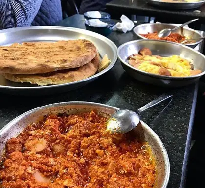 میز غذای سنتی و لذیذ ایرانی در قیمه سرای عمو هوشنگ شامل قیمه، املت سوسیس و نان گرم در سینی‌های فلزی.