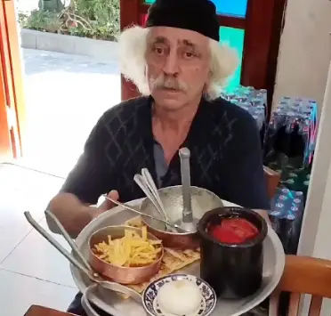 عمو هوشنگ، مردی با موهای سفید و سبیل، سینی پر از قیمه، سیب زمینی سرخ کرده و ماست را در رستوران سنتی خود حمل می‌کند.