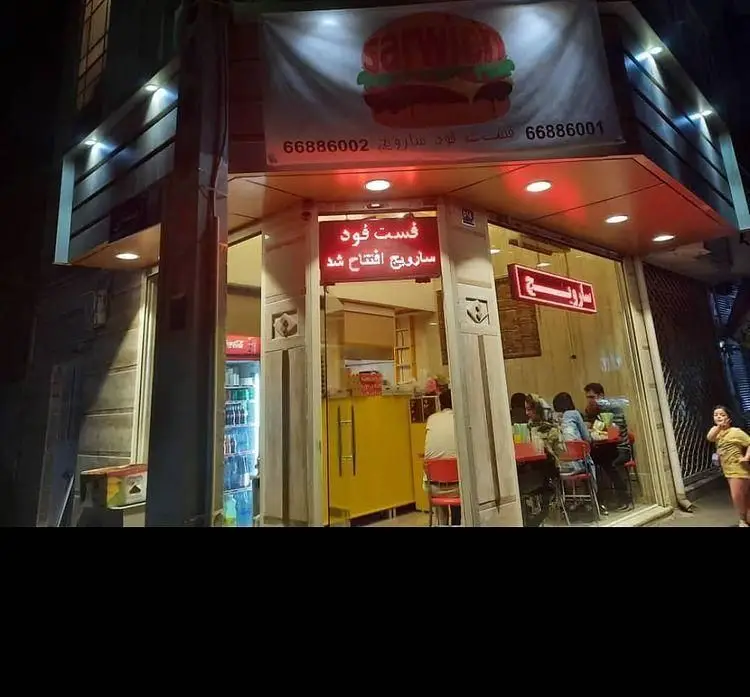 نمای شب فست فود سارویچ با تابلوی بزرگ همبرگر، نشان‌دهنده افتتاح رستوران و مشتریان داخل. شماره تماس‌ها: ۶۶۸۸۶۰۰۱، ۶۶۸۸۶۰۰۲.