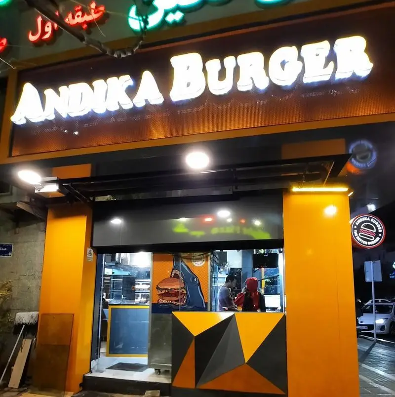 نمای بیرونی جذاب فست فود اندیکا برگر در شب با تابلوی روشن Andika Burger، دکوراسیون نارنجی و طرح کوسه در حال خوردن برگر.