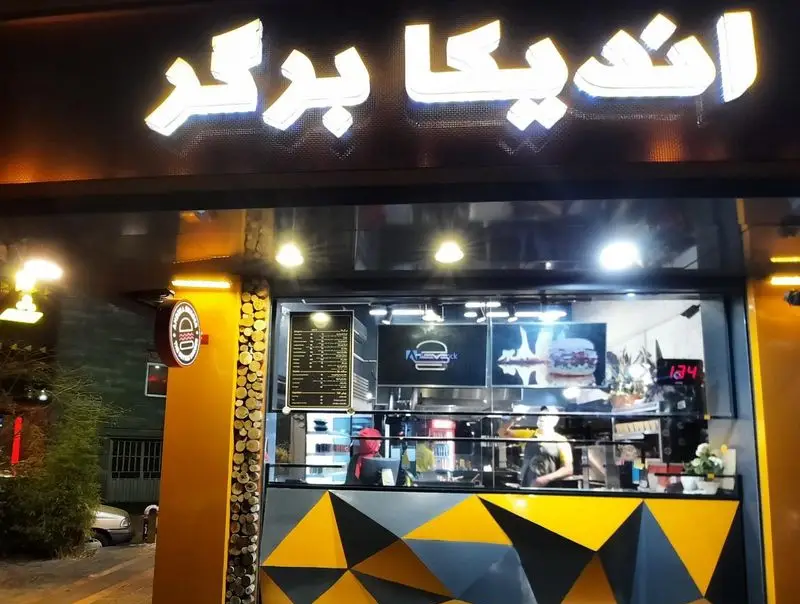 نمای شبانه فست فود اندیکا برگر با تابلوی روشن، دکوراسیون مدرن زرد و سیاه و ویترین غذاهای خوشمزه که مشتریان را جذب می‌کند.