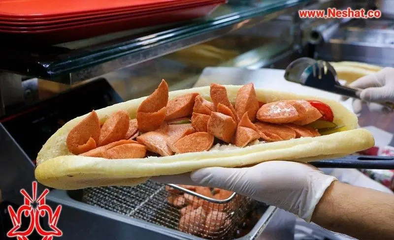 آماده سازی ساندویچ سوسیس سرخ شده بزرگ در رستوران نشاط، با نان باگت، خیارشور و گوجه فرنگی. غذای فست فود لذیذ.