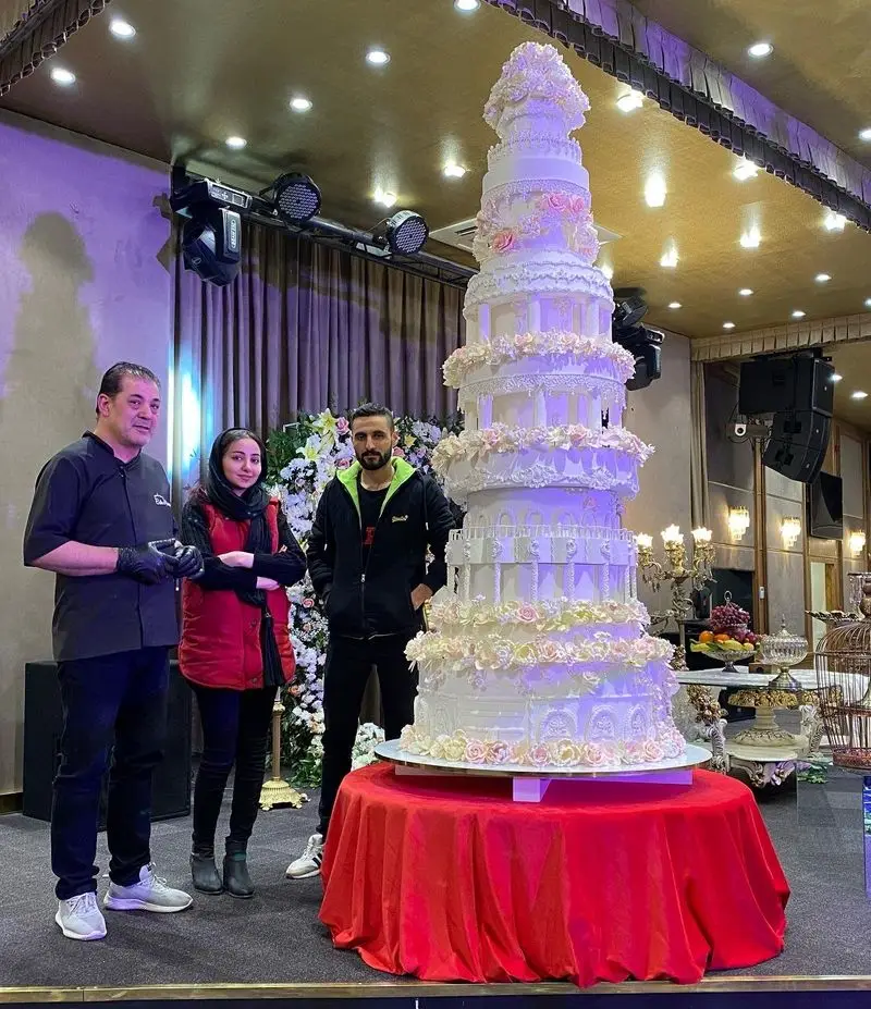 کیک عروسی عظیم و چند طبقه از کیک جواد خدادادی تبریز، تزئین شده با گل‌های زیبا، کنار تیم سازنده در مراسمی باشکوه.