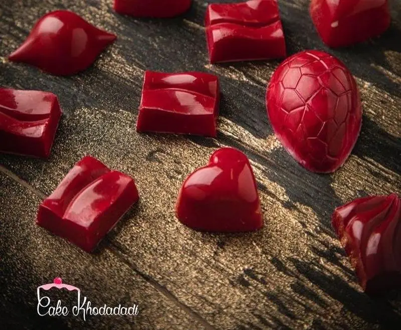 انواع شکلات‌های قرمز براق دست‌ساز کیک جواد خدادادی تبریز با طرح‌های قلب، مربع و تخم‌مرغی مناسب هدیه و ولنتاین روی پس‌زمینه تیره.