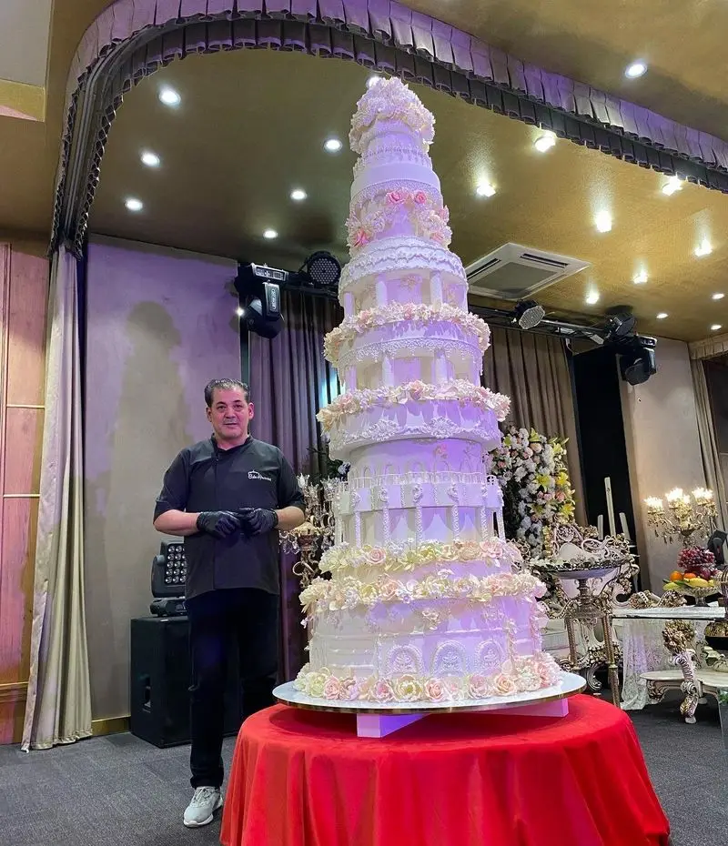 کیک عروسی دست‌ساز جواد خدادادی تبریز، بلند و مجلل با جزئیات دقیق و تزئینات گلدار در فضای شیک تالار.