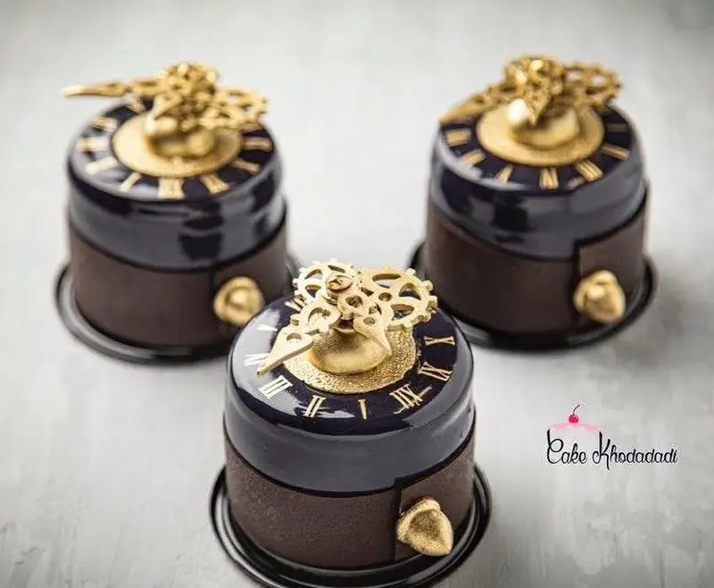 سه مینی کیک مدرن و لوکس با طرح خلاقانه ساعت رومیزی، دارای جزئیات طلایی، چرخ‌دنده و اعداد رومی، از کیک جواد خدادادی تبریز.