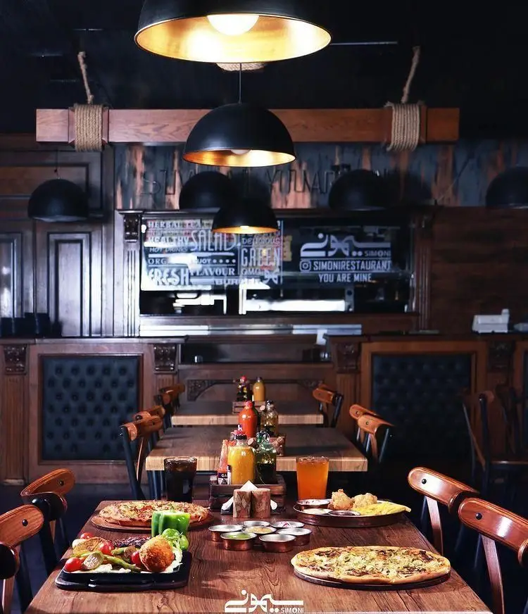 فضای داخلی کافه رستوران سیمونی، محیطی دنج و گرم با دکوراسیون چوبی، نورپردازی هنری، میزهای چیده شده با پیتزا، غذا و نوشیدنی‌های متنوع.