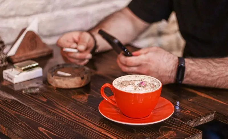 تصویری از مشتری در کافه فست فود برج برگر، در حال لذت بردن از قهوه و استفاده از موبایل، با محیطی آرام و چوبی.