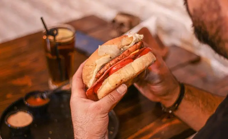 برگر آبدار با نان تازه، مرغ گریل شده، گوجه و کاهو در دست مشتری کافه فست فود برج برگر همراه با نوشیدنی و سس در پس‌زمینه.