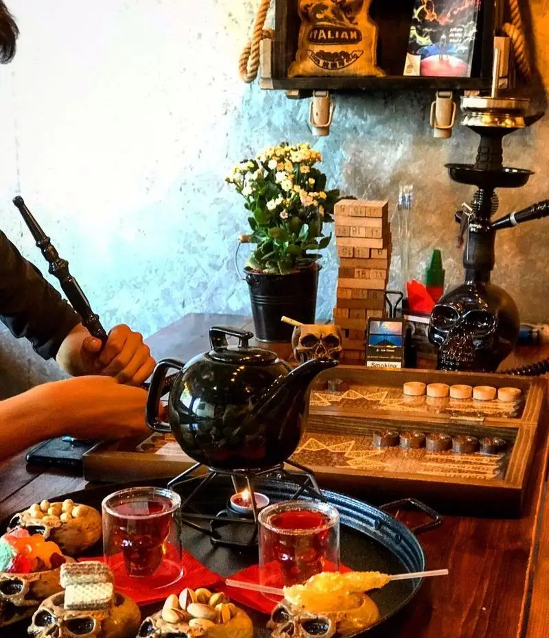 تصویری از کافه فست فود برج برگر: میز شلوغ با قلیان جمجمه‌ای، چای گرم، اسنک‌های جذاب و تخته نرد برای مشتریان.