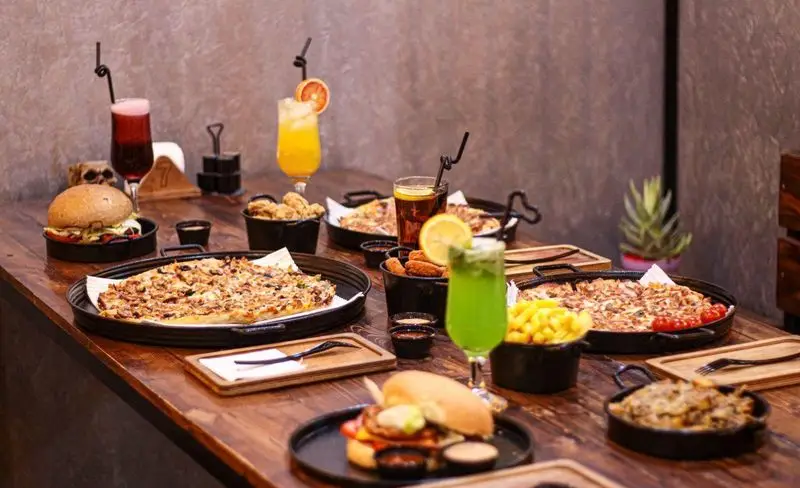 نمایی از میز پر از غذا و نوشیدنی‌های متنوع در کافه فست فود برج برگر شامل برگر، پیتزا، سیب‌زمینی و مرغ سوخاری.