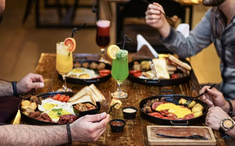 نمای نزدیک از میز پر از غذا در کافه فست فود برج برگر، شامل صبحانه مفصل با تخم مرغ، سوسیس، سیب‌زمینی، نان تست و نوشیدنی‌های رنگارنگ.