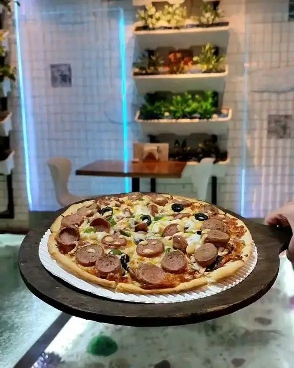 پیتزا سوسیس و زیتون در کافه رستوران آکوا با فضای داخلی مدرن، دیوار آجری سفید، نورپردازی آبی و قفسه‌های گیاهی.