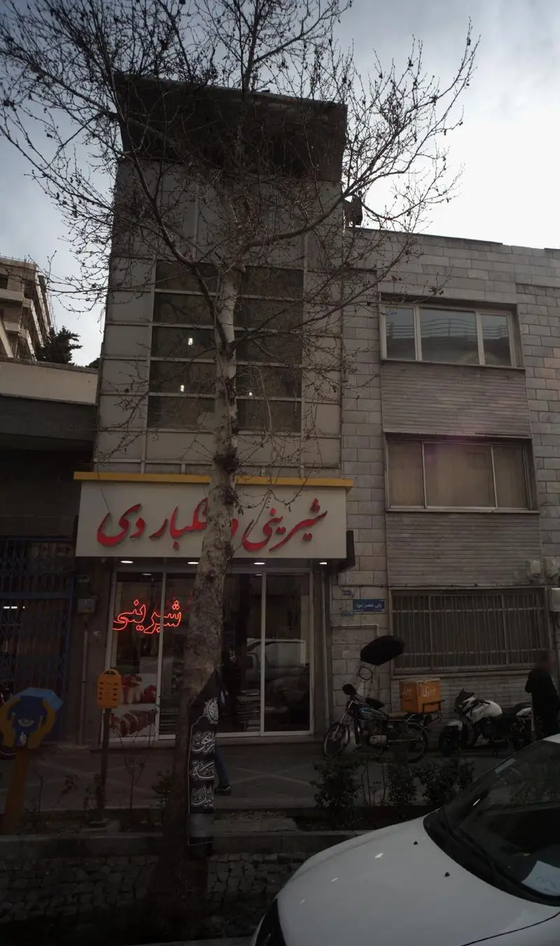 تصویری از نمای بیرونی شیرینی و خشکبار دی در خیابان ولیعصر، با تابلوی اصلی و درخت جلو مغازه. فروشگاه شیرینی و خشکبار.