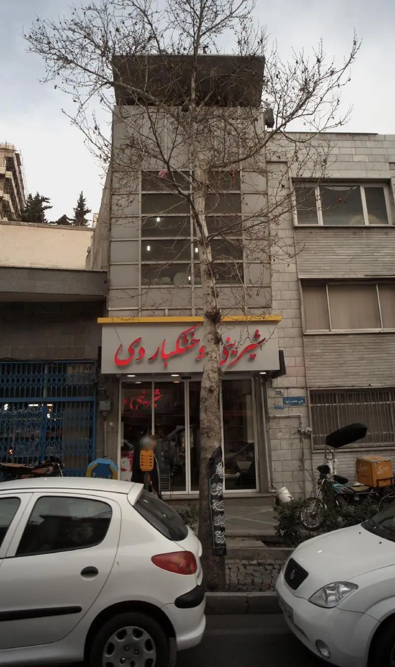 نمای بیرونی فروشگاه شیرینی و خشکبار دی در خیابان، با تابلوی مشخص و درخت بزرگ جلوی ورودی. قنادی و آجیل فروشی در شهر.