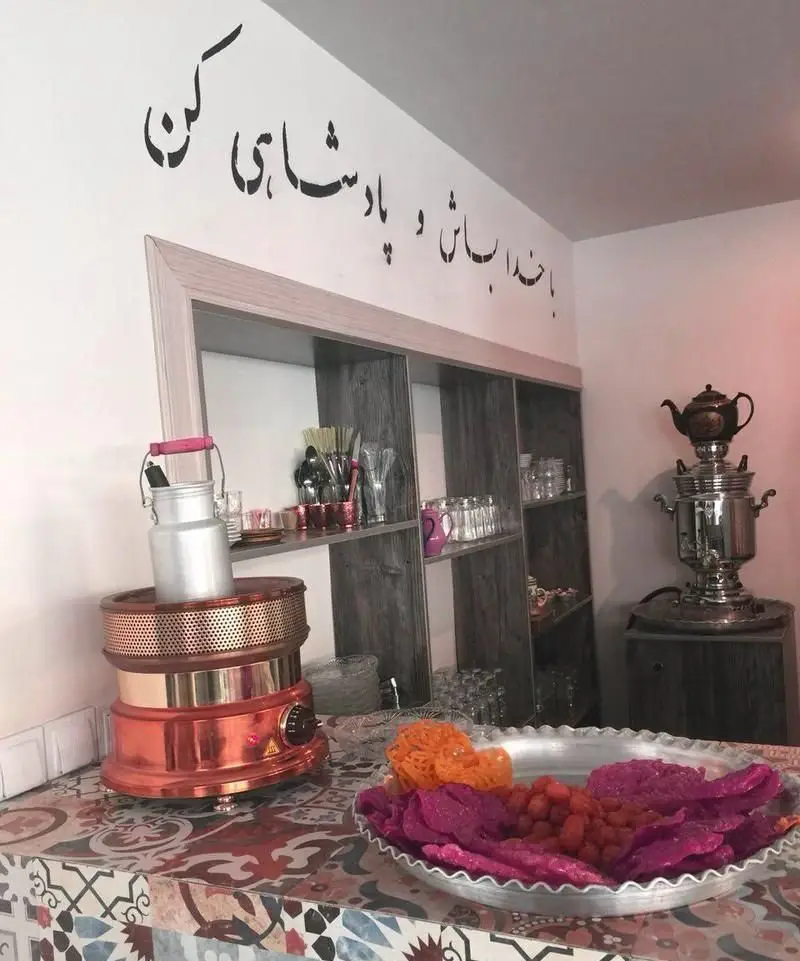 تصویری از دکوراسیون گرم و سنتی کافه رستوران شاهی، با سماور چای، شیرینی‌های رنگارنگ و خطاطی زیبای "با خدا باش و پادشاهی کن".