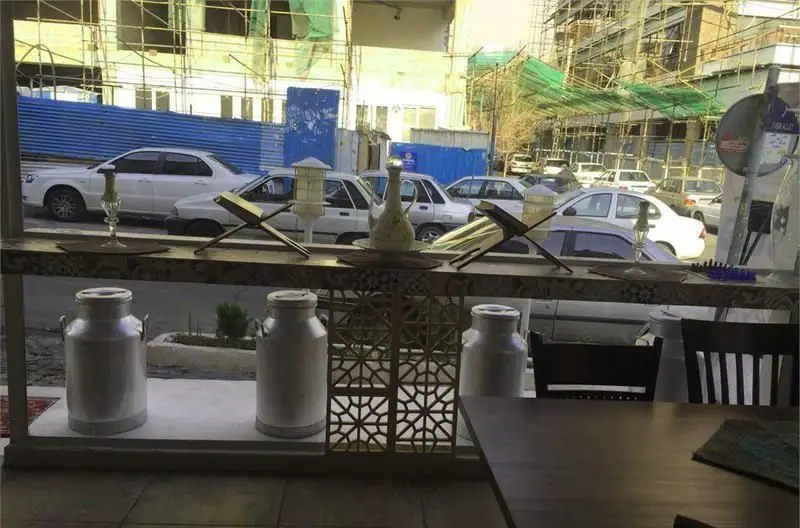 کافه رستوران شاهی: نمایی جذاب از فضای داخلی با رحل، پارچ سنتی و گالن‌های شیر، رو به خیابان پرتردد شهری و ساختمان‌های در حال ساخت.