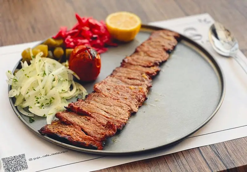 تصویری از کباب برگ لذیذ و برش خورده با دورچین پیاز و جعفری، گوجه کبابی، ترشی و لیمو در کافه رستوران گجا