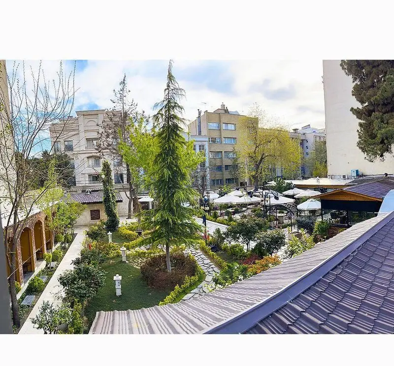 نمایی از باغ دلنشین کافه رستوران گجا با فضای سبز، میزهای چیده شده در فضای باز و درختان بلند در محیط شهری زیبا.