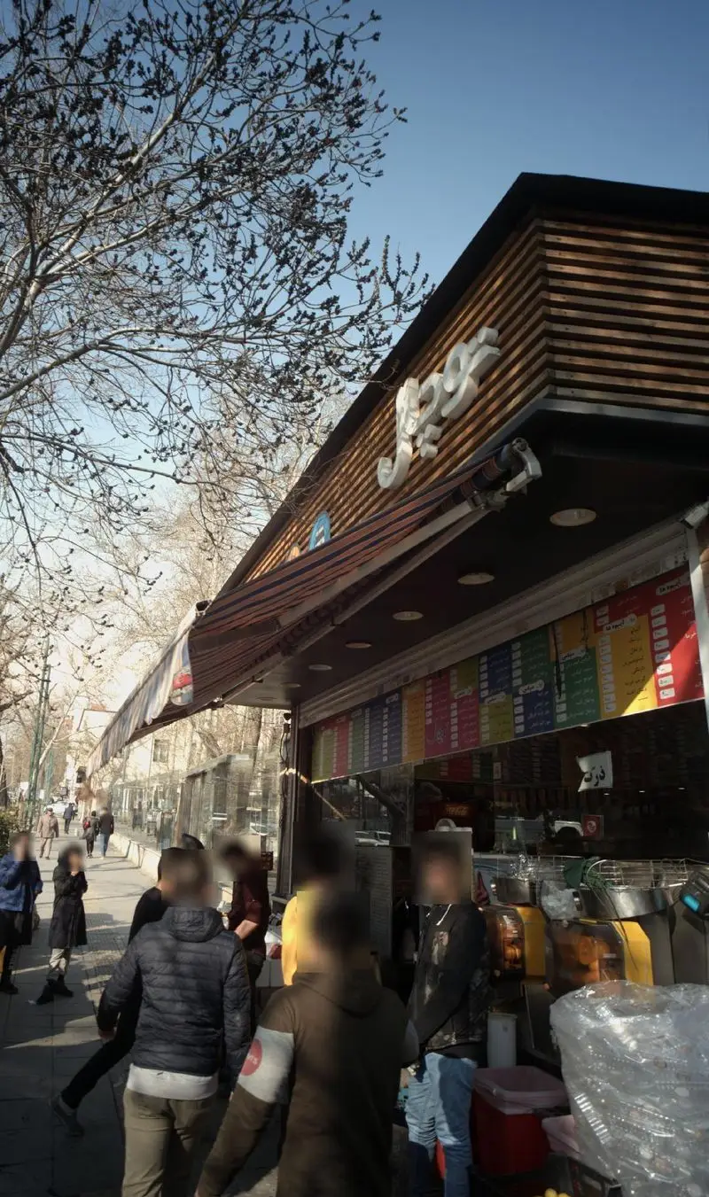 نمای خارجی آب میوه طبیعی توچال با طراحی چوبی، منوی رنگارنگ و حضور مشتریان در پیاده‌رو، محیطی دلپذیر برای خرید آبمیوه تازه.