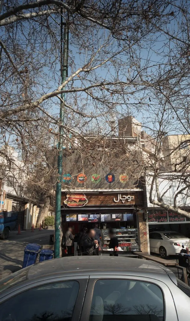 نمای بیرونی فروشگاه آب میوه طبیعی توچال، با تابلوی همبرگر و منوی فست فود، در خیابانی پر درخت و شلوغ با حضور مشتریان.