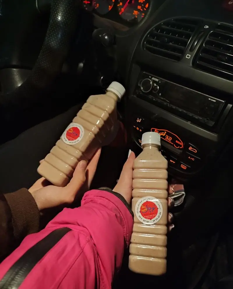 صحنه‌ای دلنشین از دو بطری آب میوه طبیعی توچال در دستان افراد داخل خودرو، با پس‌زمینه داشبورد روشن در تاریکی شب، لذت واقعی.