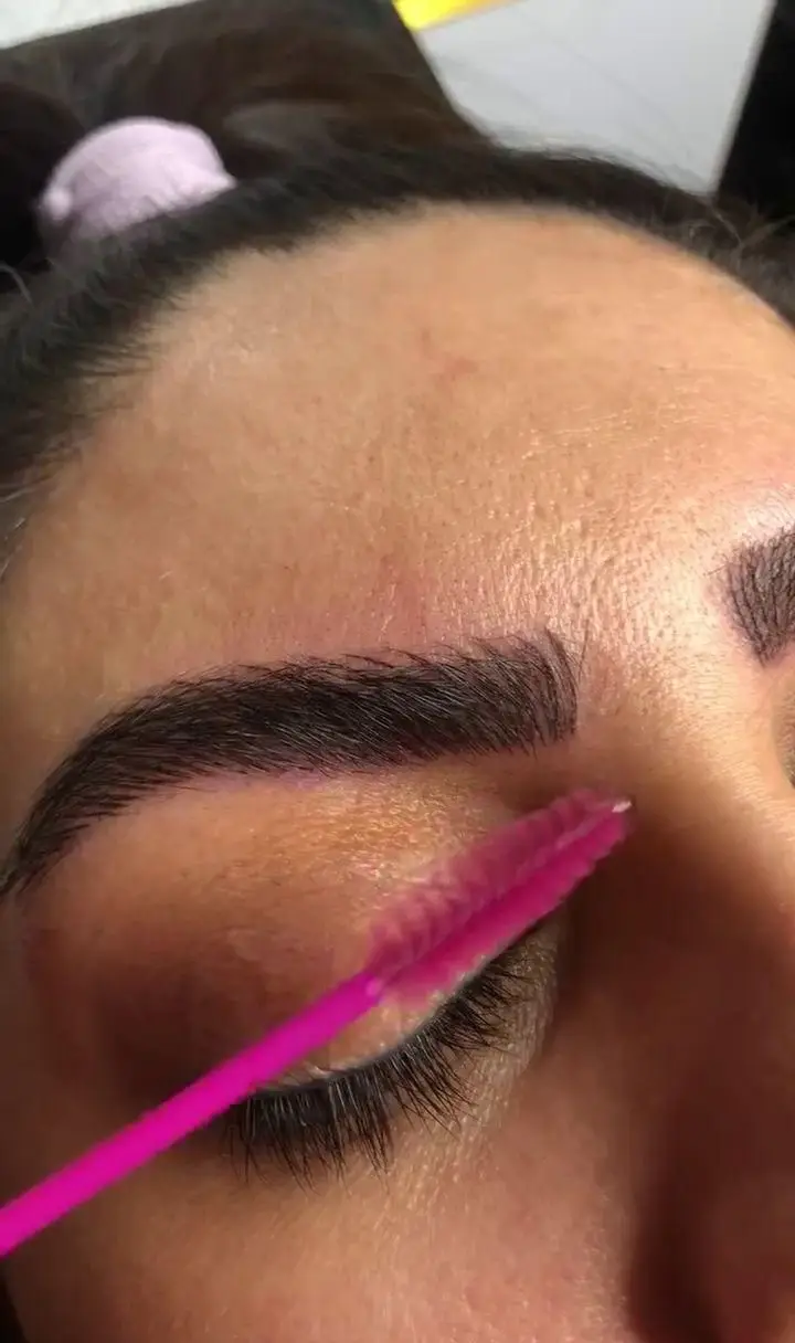 نمای نزدیک از صورت زنی که ابروهای پرپشت و مرتبش با یک برس صورتی مخصوص ابرو در حال شانه شدن است.