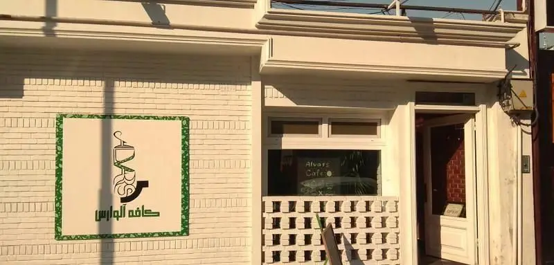 نمای ورودی کافی شاپ آلوارس با تابلوی سبز و سفید شامل نام کافه و لوگوی فنجان، درب و پنجره سفید. محیطی دلنشین و شیک برای قهوه دوستان.