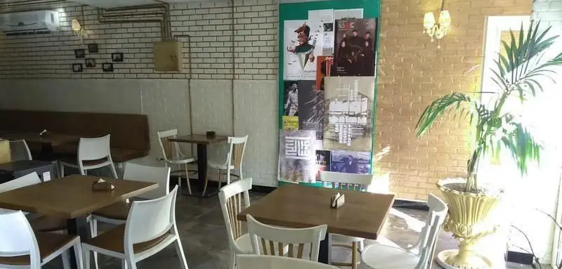 فضای دنج کافی شاپ آلوارس با دیوارهای آجری سفید و زرد، میز و صندلی چوبی، نیمکت قهوه‌ای و یک گیاه آپارتمانی بزرگ.