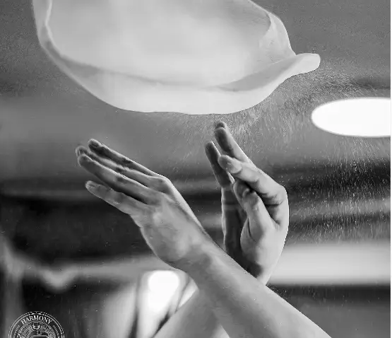 تصویری هنری از آماده‌سازی حرفه‌ای پیتزا: دستان آشپز ماهر در حال پرتاب خمیر پیتزا و آرد در آشپزخانه هارمونی.