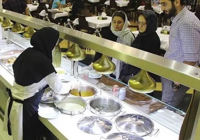 یک خانم در حال سرو غذاهای سنتی و گرم ایرانی در بوفه سلف سرویس رستوران اسلیمی برای مشتریان مشتاق.