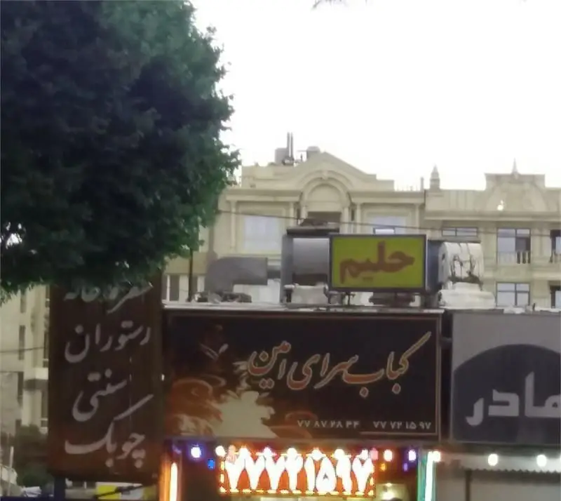 نمایی از سردر و تابلوی کباب سرای امین، با تابلوی حلیم و سایر فروشگاه‌ها در خیابانی شلوغ، همراه با درخت و ساختمان‌های شهر.