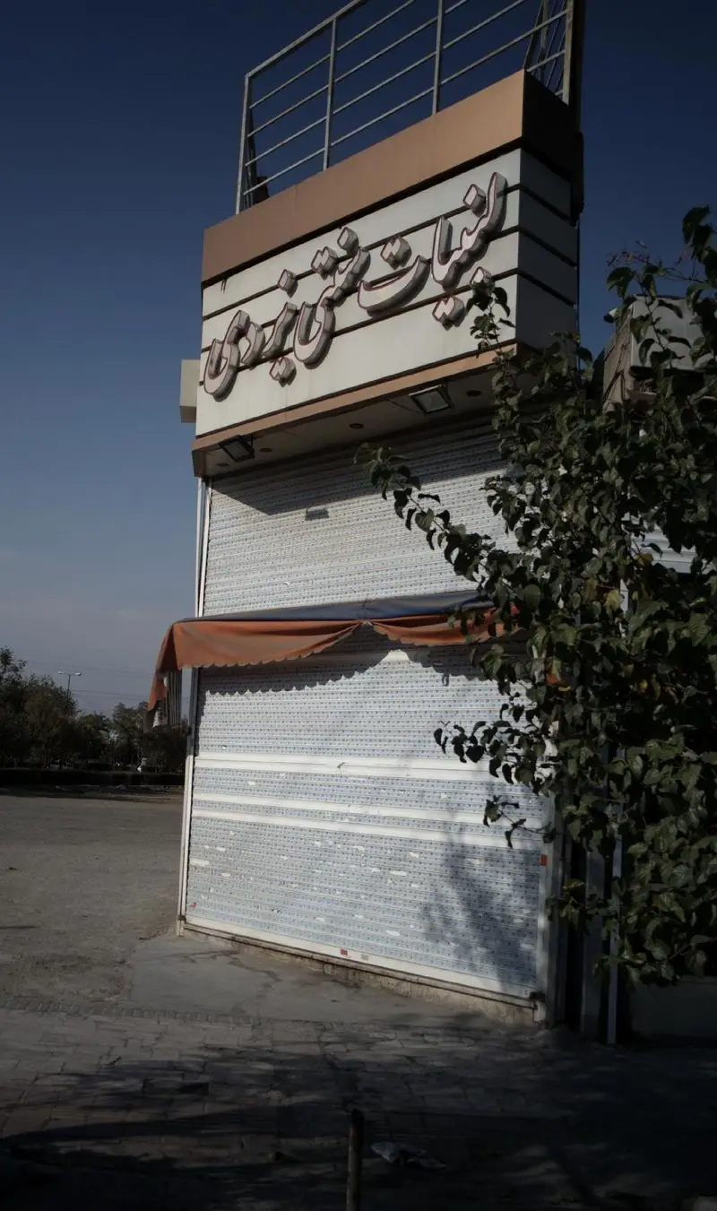 نمای خارجی مغازه لبنیات سنتی یزدی با تابلوی برجسته، کرکره‌های بسته، سایبان قهوه‌ای و درخت در کنارش. کسب و کار محصولات لبنی.