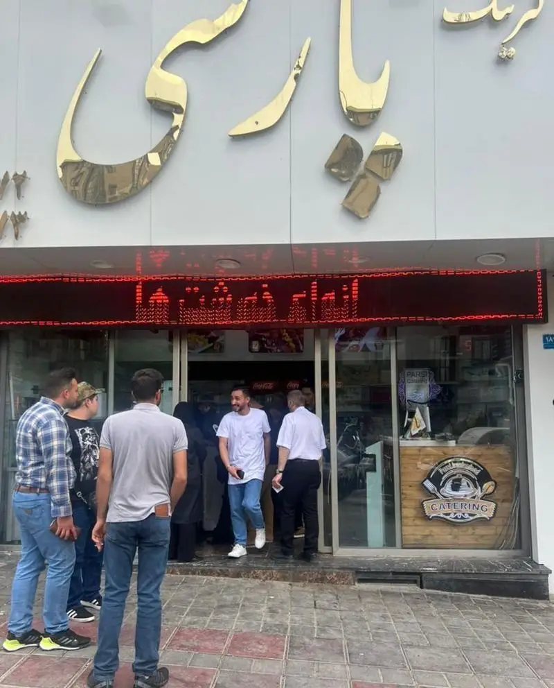 تصویر نمای بیرونی شلوغ و پر جنب و جوش کترینگ پارسی، کسب و کار تهیه غذا، با مشتریان در حال ورود و خروج و تابلوهای طلایی و LED.