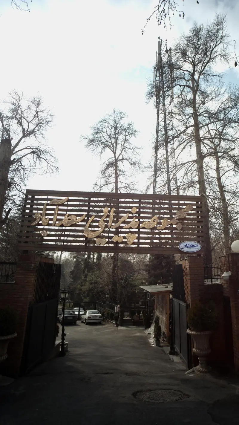 تصویری از ورودی مجموعه پذیرایی سعد آباد در تهران، با تابلوی طلایی رنگ و درختان پاییزی بلند در پس زمینه.