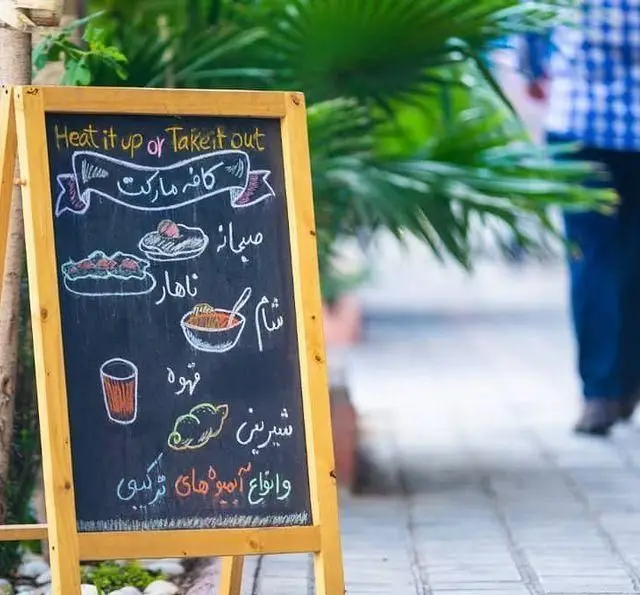 تصویری از تابلوی گچی کافه مارکت که منوی صبحانه، ناهار، شام، قهوه، شیرینی و انواع آبمیوه را نشان می‌دهد و گزینه بیرون‌بر را تبلیغ می‌کند.
