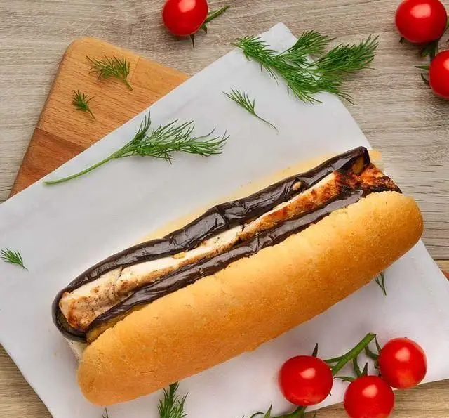 ساندویچ بادمجان گریل شده و پنیر در نان باگت، روی میز چوبی همراه با شوید و گوجه گیلاسی، یک پیشنهاد سالم از کافه مارکت.