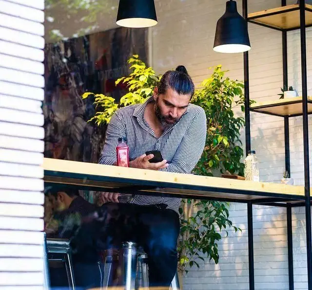 مشتری در کافه مارکت، در یک فضای آرام و مدرن، مشغول بررسی گوشی موبایل خود. دکوراسیون جذاب با میز چوبی و قفسه های فلزی.