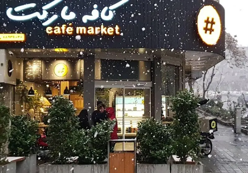 نمای بیرونی کافه مارکت در یک روز برفی زمستانی. تابلو روشن کافه مارکت (cafe market) با لوگو هشتگ، دانه های برف و اسکوتر دلیوری.