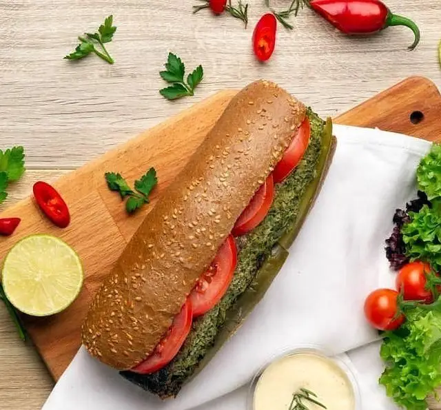 ساندویچ باگت سبزیجات تازه کافه مارکت با نان کنجدی، فیلینگ سبز، گوجه، لیمو، فلفل و سس مخصوص روی تخته چوبی، مناسب برای وعده سالم.