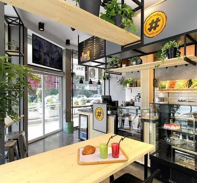 تصویری از داخل کافه مارکت با دکوراسیون شیک و مدرن، ویترین‌های متنوع کیک و دسر، نوشیدنی‌های تازه، کروسان، گیاهان سبز و فضای دنج برای مشتریان.