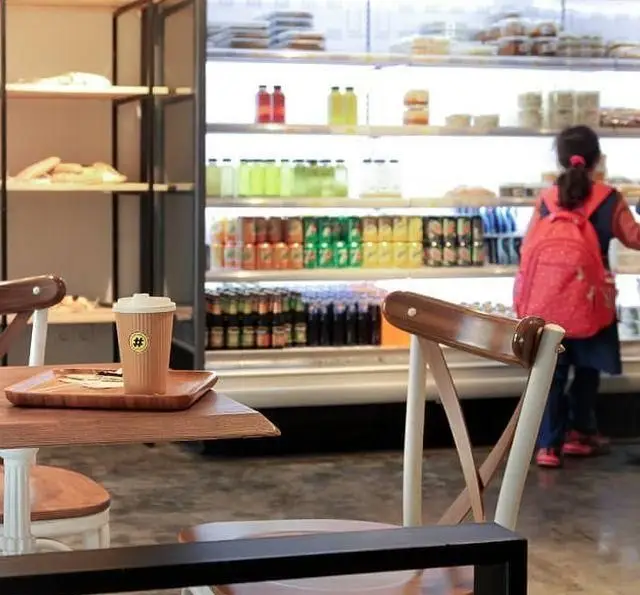 فضای داخلی کافه مارکت با قفسه‌های نان تازه، میز چوبی با فنجان قهوه بیرون‌بر و یخچال بزرگ نوشیدنی‌ها. مشتری در حال انتخاب محصولات متنوع.