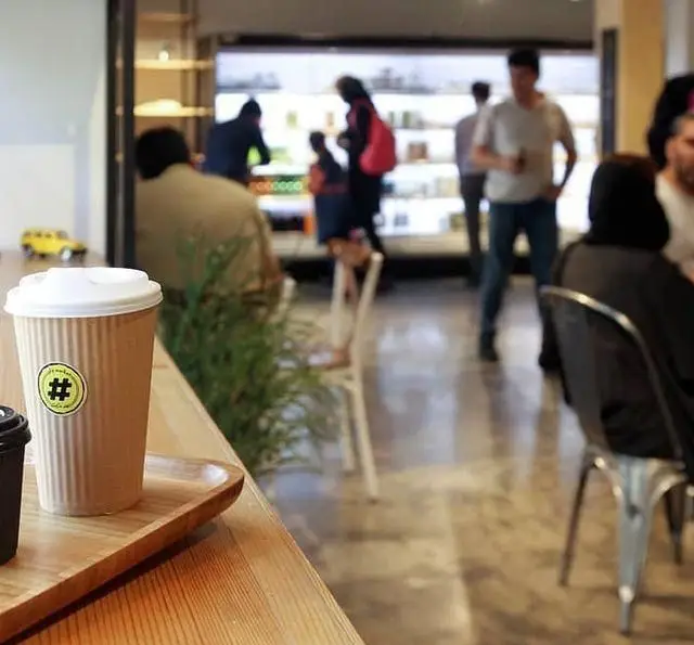 لیوان قهوه یکبار مصرف کافه مارکت با لوگوی خاص در سینی چوبی، در پیش‌زمینه شلوغ یک کافه-مارکت با مشتریان و قفسه‌های کالا.