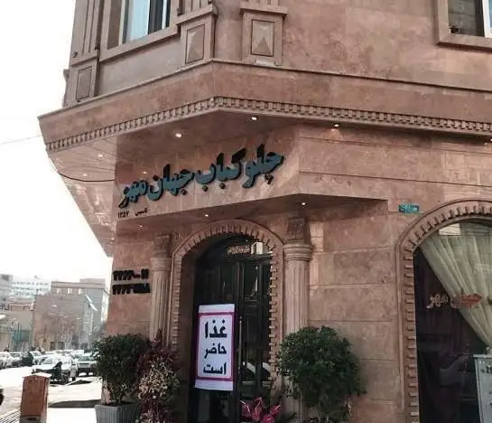 تصویر نمای بیرونی رستوران چلو کباب جهان مهر با تابلوی آبی، معماری سنگی قهوه‌ای رنگ و اعلامیه "غذا حاضر است" روی در ورودی.